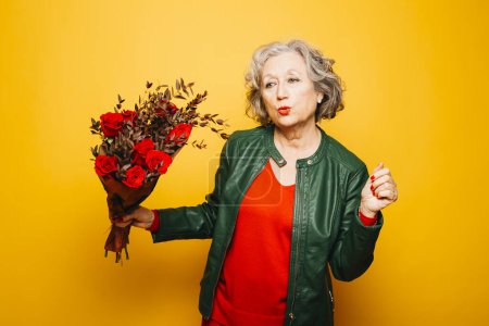 Photo for Portrait of a senior woman dancing with a red roses bouquet over a yellow background - Royalty Free Image