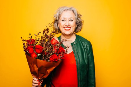 Photo for Portrait of a senior woman with a red roses bouquet over a yellow background - Royalty Free Image