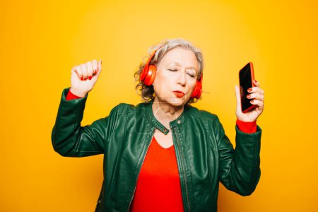 Photo for Senior woman with red headphones and red smartphone, dancing over a yellow background - Royalty Free Image