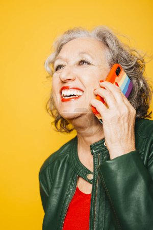 Photo for Senior woman talking on a phone with the rainbow flag on the case, over a yellow background - Royalty Free Image