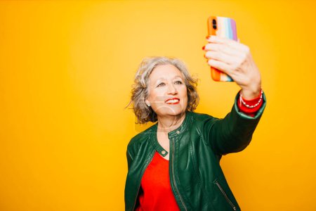 Photo for Senior woman taking a selfie with a smartphone with the rainbow flag on the case, over a yellow background - Royalty Free Image