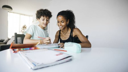 Foto de Mujer alegre ayudando a su hija a hacer la tarea. Madre feliz ayudando a su hija con la tarea escolar en la sala de estar. - Imagen libre de derechos
