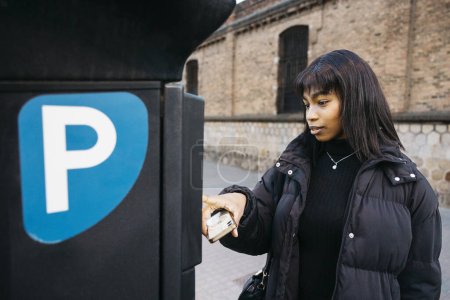 Jeune femme noire effectuant un paiement sans contact avec un smartphone au parking machine de paiement dans la rue de la ville.