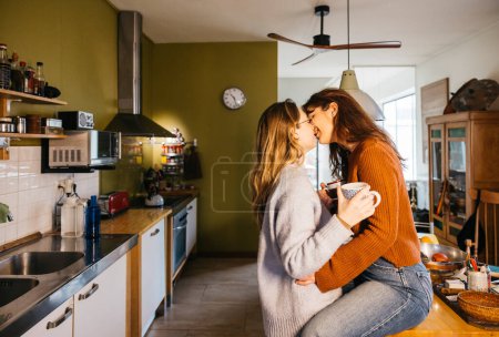 Junges weibliches Paar küsst sich bei einer Tasse Tee in der heimischen Küche. Lesbisches Paar genießt einen Moment zusammen, nippt an Tee und küsst sich in der heimischen Küche.
