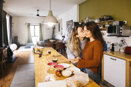 Jeune couple de femmes embrasser tout en préparant le petit déjeuner dans leur cuisine maison. Un couple de lesbiennes partage un moment intime ensemble, savourant du thé et échangeant des baisers dans l'atmosphère chaleureuse de leur cuisine à la maison.