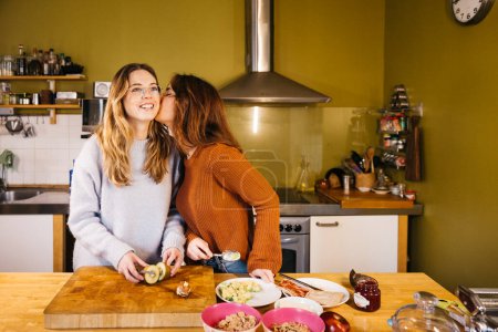 Jeune couple de femmes embrasser tout en préparant le petit déjeuner dans leur cuisine maison. Un couple de lesbiennes partage un moment intime ensemble, savourant du thé et échangeant des baisers dans l'atmosphère chaleureuse de leur cuisine à la maison.