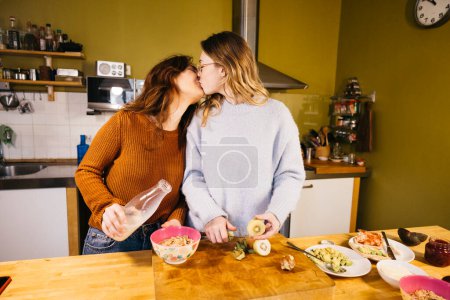 Young female couple kissing while preparing breakfast in their home kitchen. A lesbian couple shares an intimate moment together, savoring tea and exchanging kisses in the cozy atmosphere of their kitchen at home.