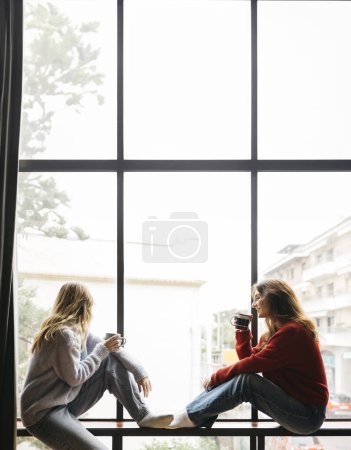 Zwei junge Frauen sitzen am Fenster und trinken Tee, in ihrem Haus. Junges Paar verbringt Zeit miteinander in intimer und gemütlicher Atmosphäre.