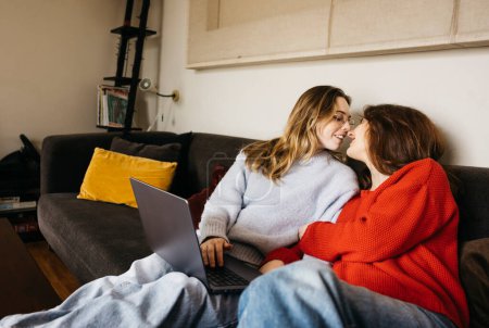 Lesbisches Paar sitzt auf einem Sofa mit einem Laptop und küsst sich. Junge schwule Paar mit einem Laptop in einer intimen und gemütlichen Atmosphäre.