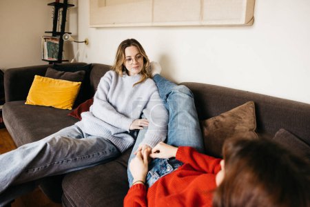 Lesbisches Paar liegt zu Hause auf dem Sofa, spricht leise und streichelt sich gegenseitig. Junge Frauen teilen einen Moment der Zärtlichkeit und Zuneigung in einer intimen und gemütlichen Atmosphäre.