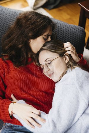 Lesbisches Paar liegt auf dem Wohnzimmerboden, umarmt und streichelt sich. Junge Frauen teilen einen Moment der Zärtlichkeit und Zuneigung in einer intimen und gemütlichen Atmosphäre.
