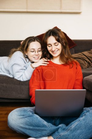 Lesbisches lächelndes Paar, das zu Hause mit einem Laptop auf einem Sofa sitzt. Junge schwule Frauen haben Spaß mit einem Laptop in einer intimen und gemütlichen Atmosphäre.
