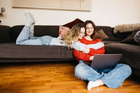 Lesbisches Paar liegt zu Hause auf dem Sofa, spricht leise und streichelt sich mit einem Laptop. Junge Frauen teilen einen Moment der Zärtlichkeit und Zuneigung in einer intimen und gemütlichen Atmosphäre.