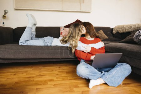 Lesbisches Paar küsst sich und benutzt einen Laptop, während es zu Hause auf einem Sofa liegt. Junges weibliches Paar küsst und streichelt sich, während es zu Hause auf einem Laptop im Internet surft.