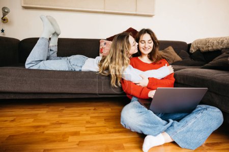 Lesbisches Paar liegt zu Hause auf dem Sofa, spricht leise und streichelt sich mit einem Laptop. Junge Frauen teilen einen Moment der Zärtlichkeit und Zuneigung in einer intimen und gemütlichen Atmosphäre.