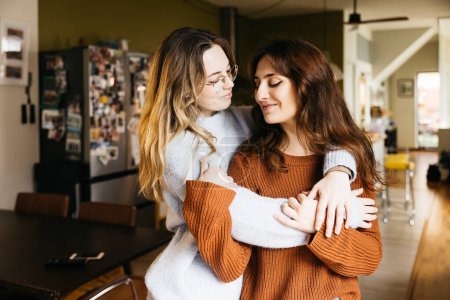 Tierno retrato de una joven hermosa pareja de lesbianas abrazándose en la acogedora sala de estar de su casa.