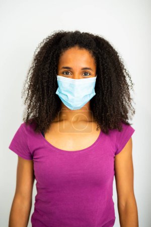 Portrait of a woman with afro hair wearing a blue surgical mask and a purple shirt, standing against a white plain background.