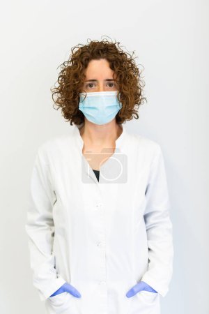 Portrait of a healthcare worker wearing a blue protective mask. Doctor wearing a white coat standing against a white backgroud and looking directly at the camera.