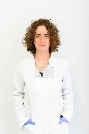 Portrait of a smiling healthcare worker wearing a white coat standing against a white backgroud and looking directly at the camera.