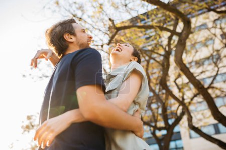 Téléchargez les photos : Jeune couple embrassant à l'extérieur, riant ensemble sous les arbres avec la lumière du soleil filtrant à travers les branches. - en image libre de droit