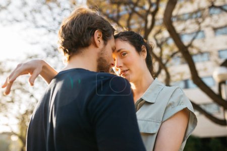 Couple embrasse étroitement, fronts touchant, dans un moment intime à l'extérieur avec des arbres et des bâtiments en arrière-plan.