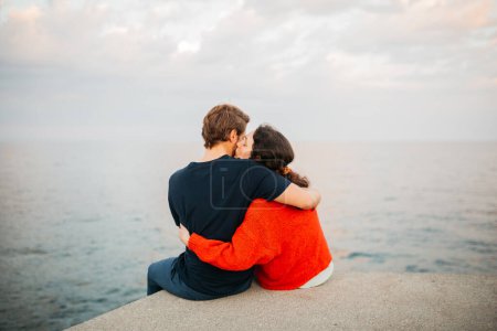 Couple assis ensemble sur un quai, embrassant et regardant au-dessus de la mer calme sous un ciel nuageux, partageant un moment paisible.