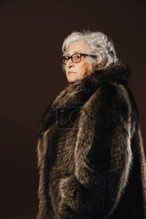 Tournage en studio d'une femme âgée sérieuse aux cheveux blancs bouclés et aux lunettes détournées debout de profil, vêtue d'un manteau de fourrure, sur un fond noir uni.