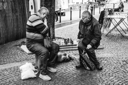 Foto de Turín, Italia. En Piazza Emanuele Filiberto, con una expresión absorbida, dos ancianos juegan al ajedrez en un banco en una pequeña plaza arbolada. Imagen en blanco y negro. 2023-05-04. - Imagen libre de derechos