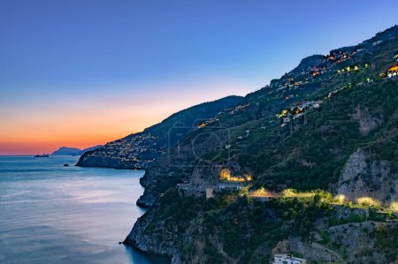 Amalfiküste, Italien. Blick über Praiano an der Amalfiküste in der Abenddämmerung. Straßen- und Hausbeleuchtung in der Abenddämmerung. In der Ferne am Horizont die Insel Capri. Amalfiküste. Meereslandschaft.