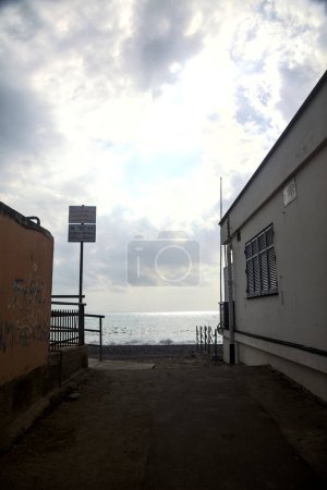 Téléchargez les photos : Accès à une plage au bord de la mer encadrée par deux bâtiments bas par une journée nuageuse - en image libre de droit