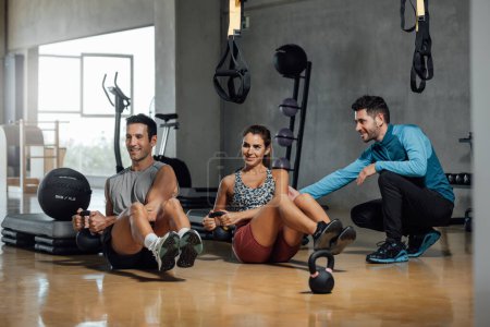 Group of smiling sporty woman and men doing russian twists abs exercises sit up, raising legs and holding kettlebell in gym. Personal trainer corrects pose for good workout. Horizontal