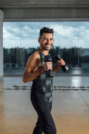 Portrait of a smiling caucasian male personal instructor, looking at camera, exercising with dumbbells in a gym. Workout lifting dumbbell weights. Vertical, window background.