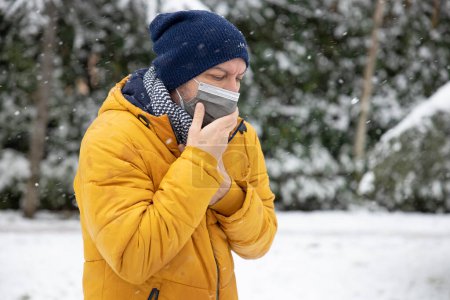 Téléchargez les photos : Sick young man with protective face mask coughing in the park on a cold snowy winter day - en image libre de droit