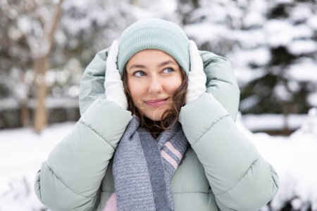 Téléchargez les photos : Jeune femme en vêtements chauds par une journée froide et enneigée. Souriant, couvrant ses oreilles avec sa main gantée - en image libre de droit