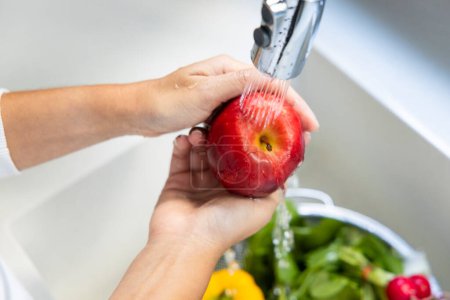 Téléchargez les photos : Femme lavant une pomme rouge dans l'évier dans la cuisine et d'autres délicieux légumes crus d'hiver dans la passoire dans l'évier - en image libre de droit
