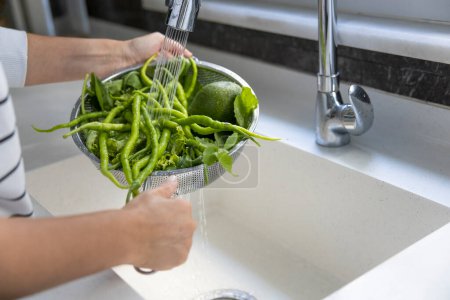 Foto de Mujer joven lavando chiles verdes picantes en colador por fregadero de cocina - Imagen libre de derechos