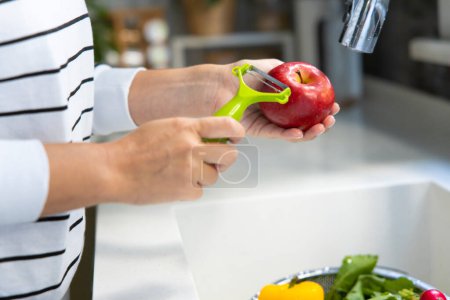 Photo for Young woman peeling an apple with peeler in kitchen and other delicious raw winter vegetables in the strainer in the sin - Royalty Free Image