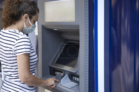 Téléchargez les photos : Jeune femme avec masque protecteur sur une banque ATM prendre de l'argent - en image libre de droit