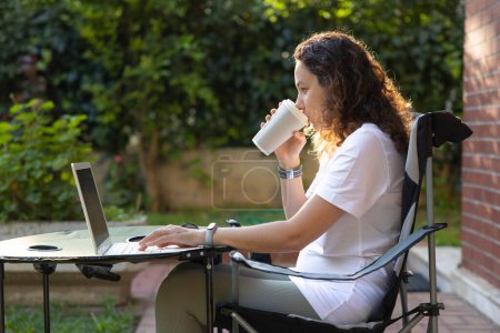 Téléchargez les photos : Jeune femme assise sur une chaise dans le jardin de la cour arrière et regardant lapto - en image libre de droit