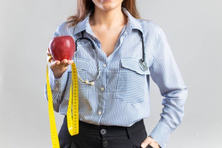 Photo for Young nutritionist holding a tape measure and an apple, looking at camera and smiling on gray background - Royalty Free Image