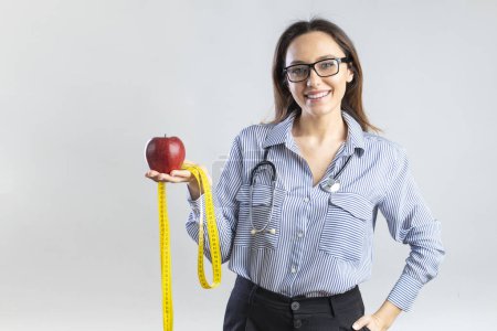 Téléchargez les photos : Jeune nutritionniste tenant un ruban à mesurer et une pomme, regardant la caméra et souriant sur fond gris - en image libre de droit