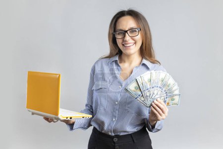 Foto de Young successful business woman making money from internet, holding cash and laptop in hands, gray background. E-commerce and profit online. - Imagen libre de derechos