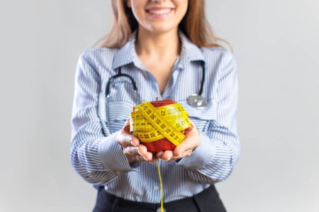 Téléchargez les photos : Jeune nutritionniste tenant un mètre ruban et une pomme, regardant la caméra et souriant sur fond gris - en image libre de droit