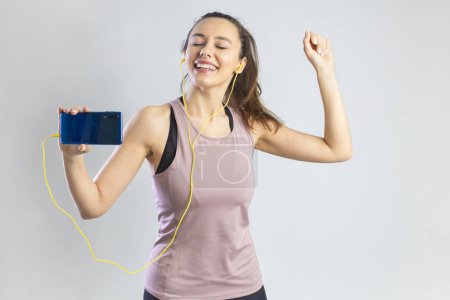 Téléchargez les photos : Jeune femme sportive dans un écouteur tenant smartphone et dansant sur fond gris - en image libre de droit