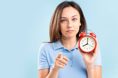 Téléchargez les photos : Young woman holding a red alarm clock while pointing at camera on blue background - en image libre de droit