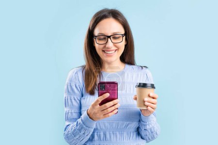 Photo for Smiling young woman with eyeglasses, holding smartphone and paper coffee cup standing isolated over blue backgroun - Royalty Free Image