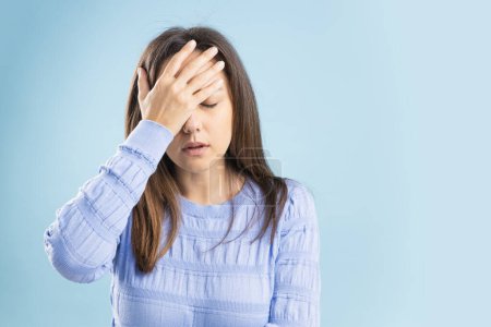 Photo for Portrait of upset, sad woman having head ache, stress, troubles, touching head and close eyes, standing over blue background - Royalty Free Image