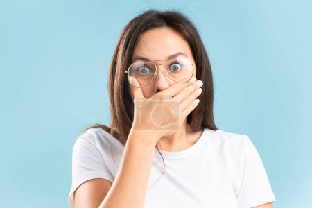Téléchargez les photos : Stunned young woman looking away with hand on mouth against blue background. Female with eyeglasses looking terrified. - en image libre de droit
