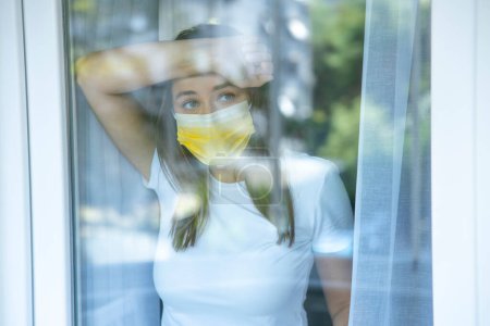 Foto de Mujer joven que no puede salir de la casa en cuarentena debido a una epidemia de Covid-19 y mirando por la ventana - Imagen libre de derechos
