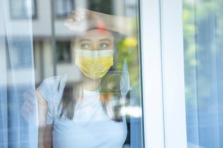 Foto de Young woman who cannot leave the house in quarantine due to an epidemic Covid-19 and looking through the window - Imagen libre de derechos